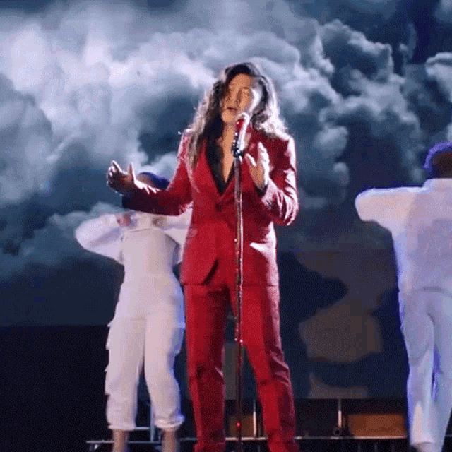 a woman in a red suit is singing into a microphone while two dancers are dancing in the background .