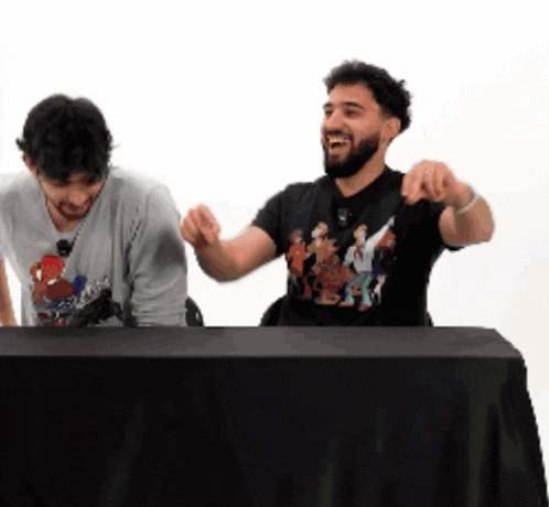 two men are sitting at a table with their hands in the air and one is wearing a shirt with a cartoon on it