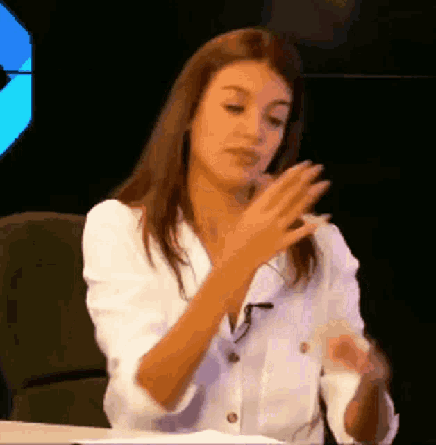 a woman in a white shirt is sitting at a table with her hands up .