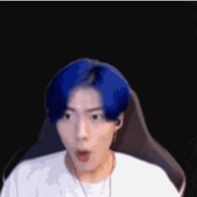 a man with blue hair is sitting in a chair making a surprised face .