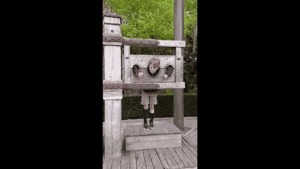 a boy is standing in a wooden stockade with his head in a hole .