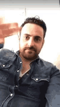 a man with a beard is taking a selfie with his phone while wearing a denim shirt .