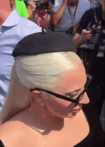 a blonde woman wearing glasses and a black hat stands in a crowd