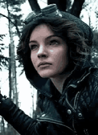 a woman in a hooded jacket and goggles is standing in a forest .