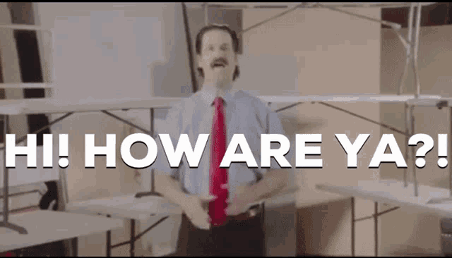 a man in a blue shirt and red tie is standing in front of folding tables and says hi how are ya
