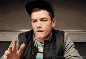 a young man wearing a hat and a vest is making a gesture with his hands .