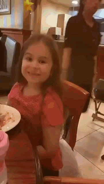 a little girl sits at a table with a plate of food in front of her