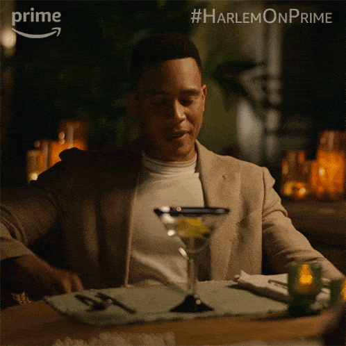 a man sits at a table with a martini in front of him and #harlemonprime written above him