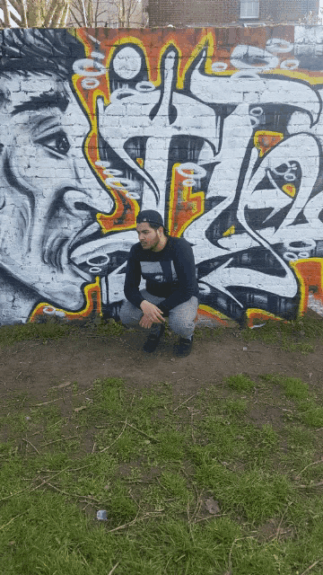 a man squatting in front of a wall with graffiti on it that says ' s '