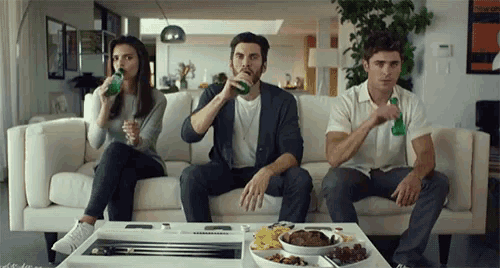 three people are sitting on a couch drinking beer and eating snacks