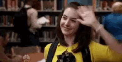 a woman in a yellow shirt is waving at the camera in a library while wearing a backpack .