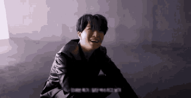 a young man in a black leather jacket is sitting on the floor and smiling .