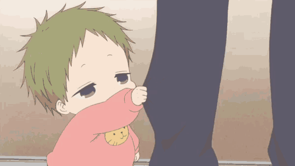 a baby with green hair is covering his face with his hand while standing next to a person 's leg .