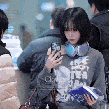 a girl wearing headphones and a sweatshirt that says clover looks at her phone