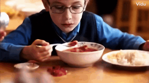a young boy wearing glasses is eating a bowl of soup with a spoon