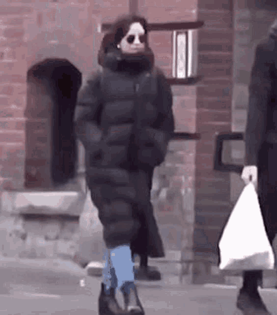 a woman in a long black coat and sunglasses is walking down the street .