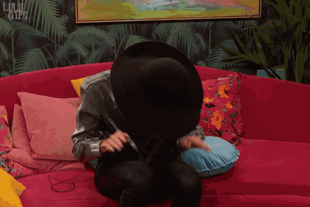 a person wearing a black hat is sitting on a pink couch with pillows