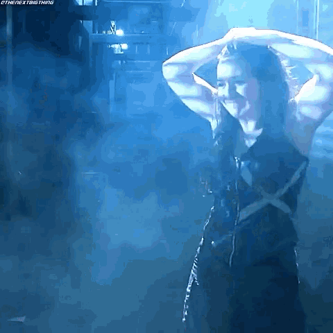 a woman in a black dress is standing in a dark room with blue smoke coming out of the ceiling .