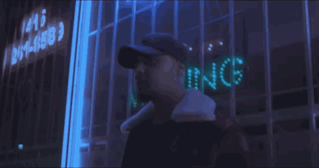 a man in a hat stands in front of a neon sign that says 1210