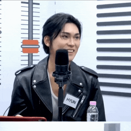a young man wearing a leather jacket and a name tag that says hyun stands in front of a microphone