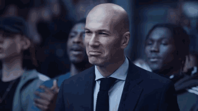 a bald man in a suit and tie stands in front of a crowd with the word that 's visible