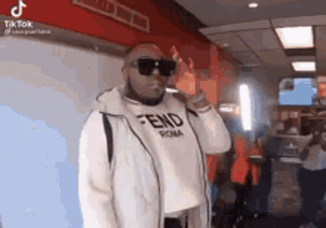 a man wearing sunglasses and a white fendi shirt is standing in a room .