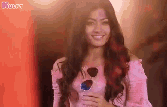 a woman wearing sunglasses and a pink off the shoulder top is smiling and looking at the camera .