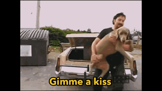 a man is holding a large dog in front of a car with the words " gimme a kiss " on the bottom