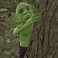 a green cartoon character standing next to a tree trunk