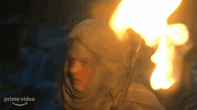a man in a hood is holding a torch in his hand and a fire is coming out of his head .