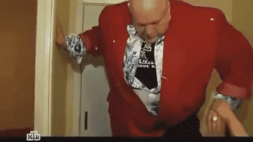 a bald man in a red jacket and tie is standing in a doorway with a tv screen behind him that says ntv