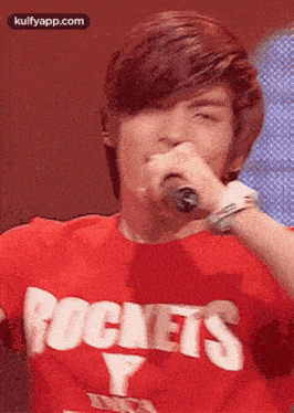 a young man wearing a red rockets t-shirt is singing into a microphone .