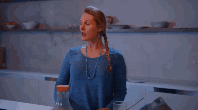 a woman in a blue sweater stands in a kitchen with a bottle and a glass