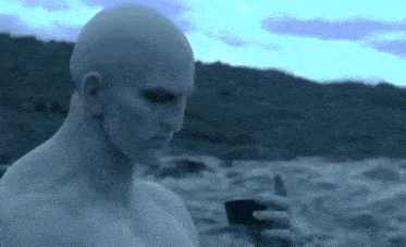 a man without a shirt is holding a cell phone while standing on a beach .