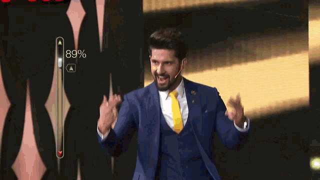 a man in a blue suit and yellow tie stands in front of a screen that shows a percentage of 89%