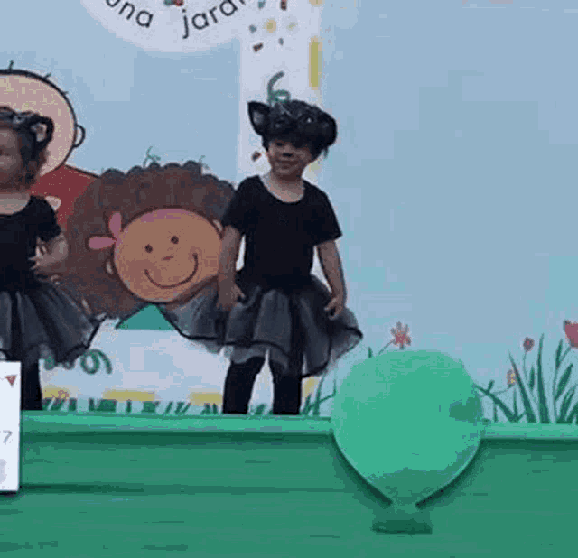two little girls are dancing on a stage with a green balloon in the background .
