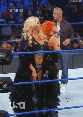 two women are standing in a wrestling ring with a man standing behind them