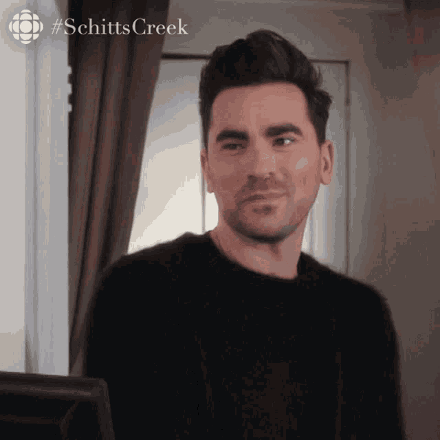 a man in a black sweater is standing in front of a computer screen with #schittscreek written on the bottom right