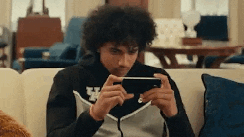 a man with curly hair is sitting on a couch playing a game on his phone .