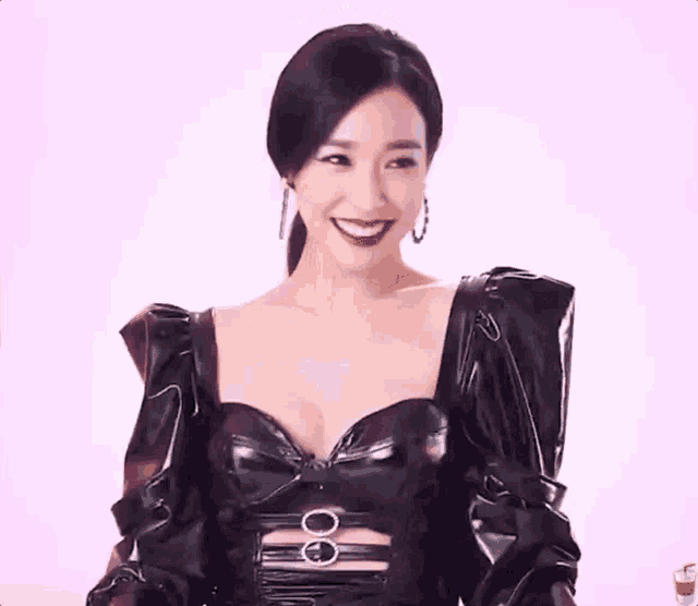 a woman in a black leather dress is smiling and looking at the camera on a pink background .
