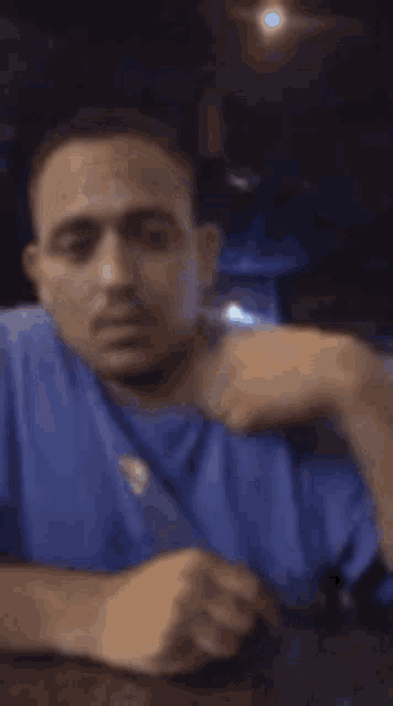 a man in a blue shirt is sitting at a table with his hand on his shoulder .