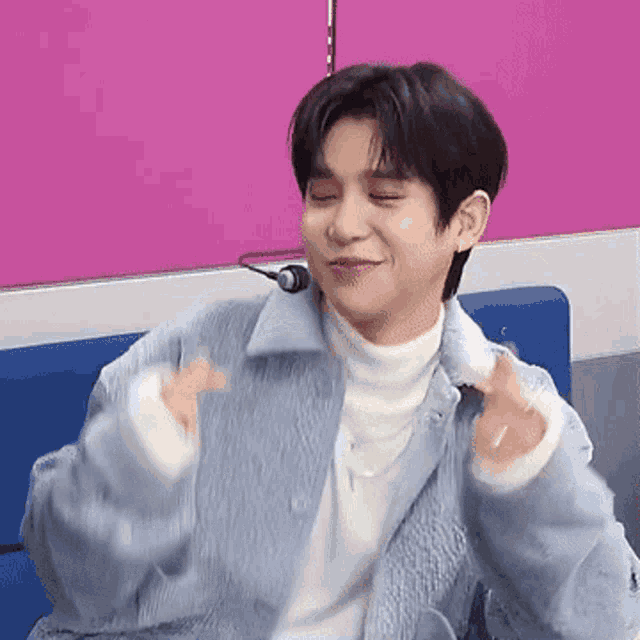 a young man is sitting on a blue couch with his eyes closed and making a heart shape with his hands .