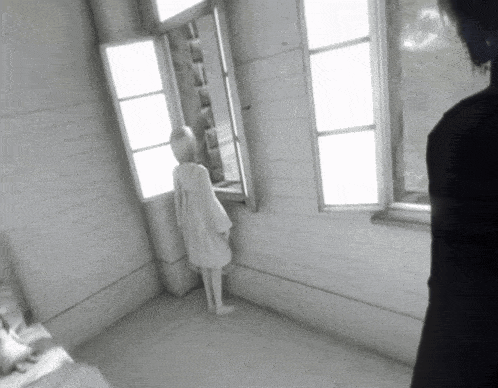 a woman in a white dress stands in a room looking out a window