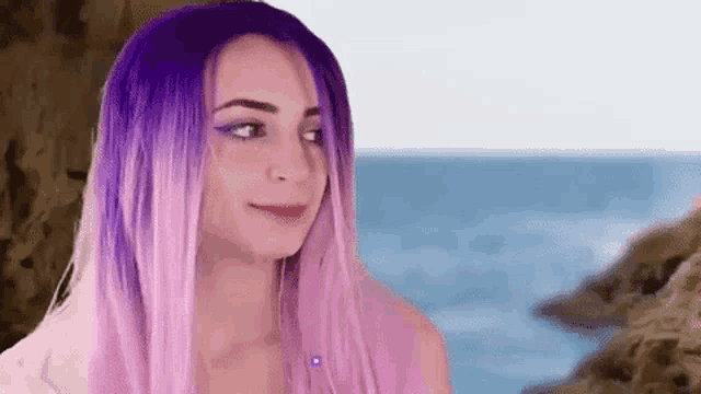 a woman with purple and pink hair is standing on a rocky beach near the ocean .