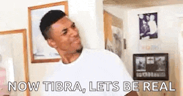 a man in a white shirt is standing in a living room and says `` now tibra , let 's be real '' .
