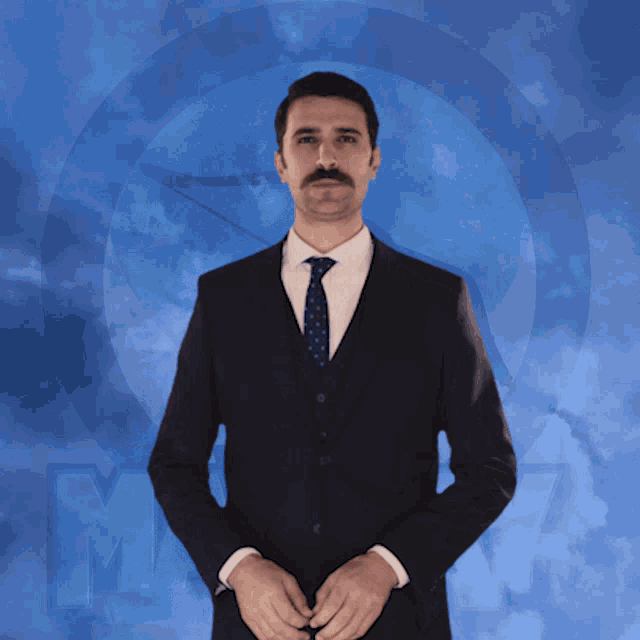 a man in a suit and tie stands in front of a blue background with the letter m visible