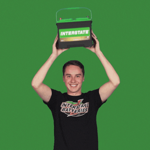 a man wearing an interstate batteries shirt is holding a green battery over his head
