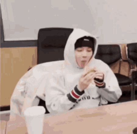 a person in a hoodie is sitting at a table eating a sandwich and drinking coffee .