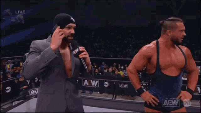 a wrestler is talking into a microphone while another wrestler stands behind him
