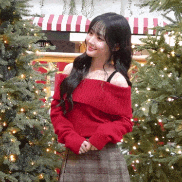 a woman in a red off the shoulder sweater is standing in front of a christmas tree